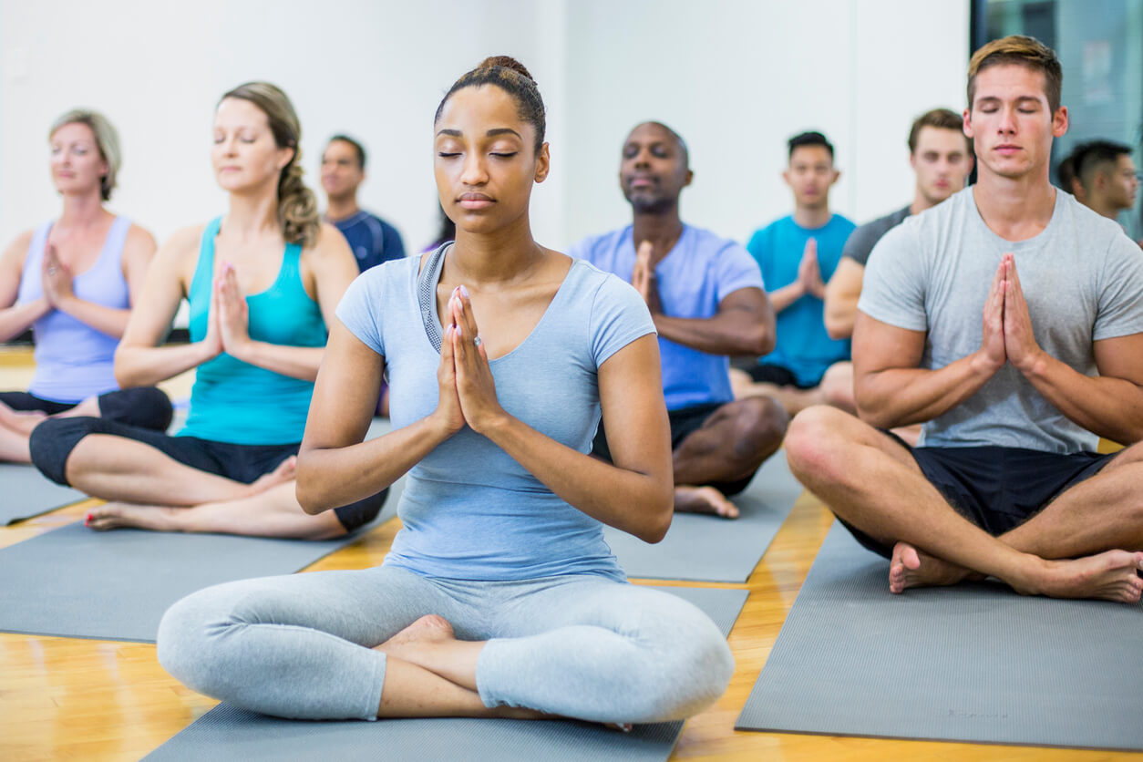 A multi-ethnic group of adults are indoors in a fitness center. They are wearing exercise clothes and shoes. Here they are doing meditation with their eyes closed.